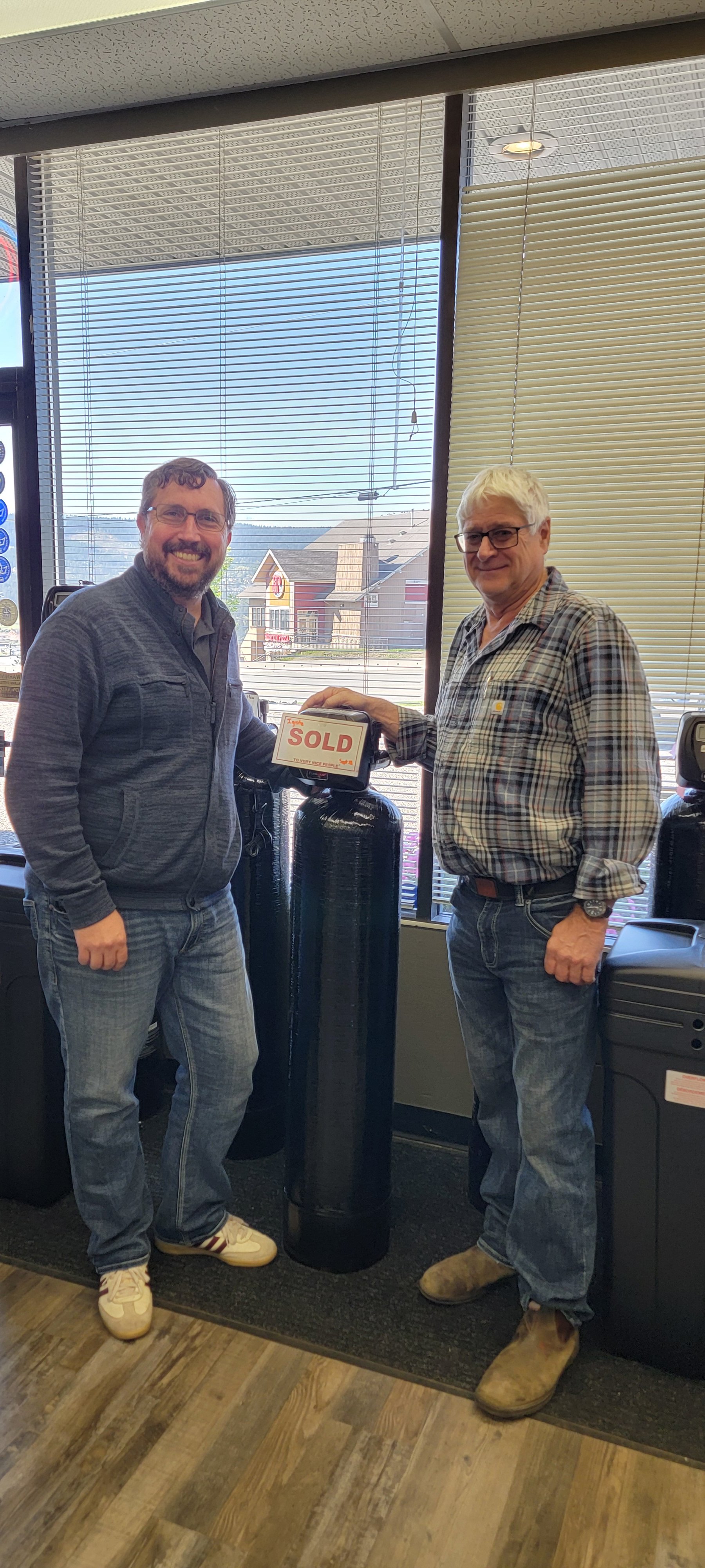 Two smiling men stand indoors beside a large case displaying a “SOLD” sign.