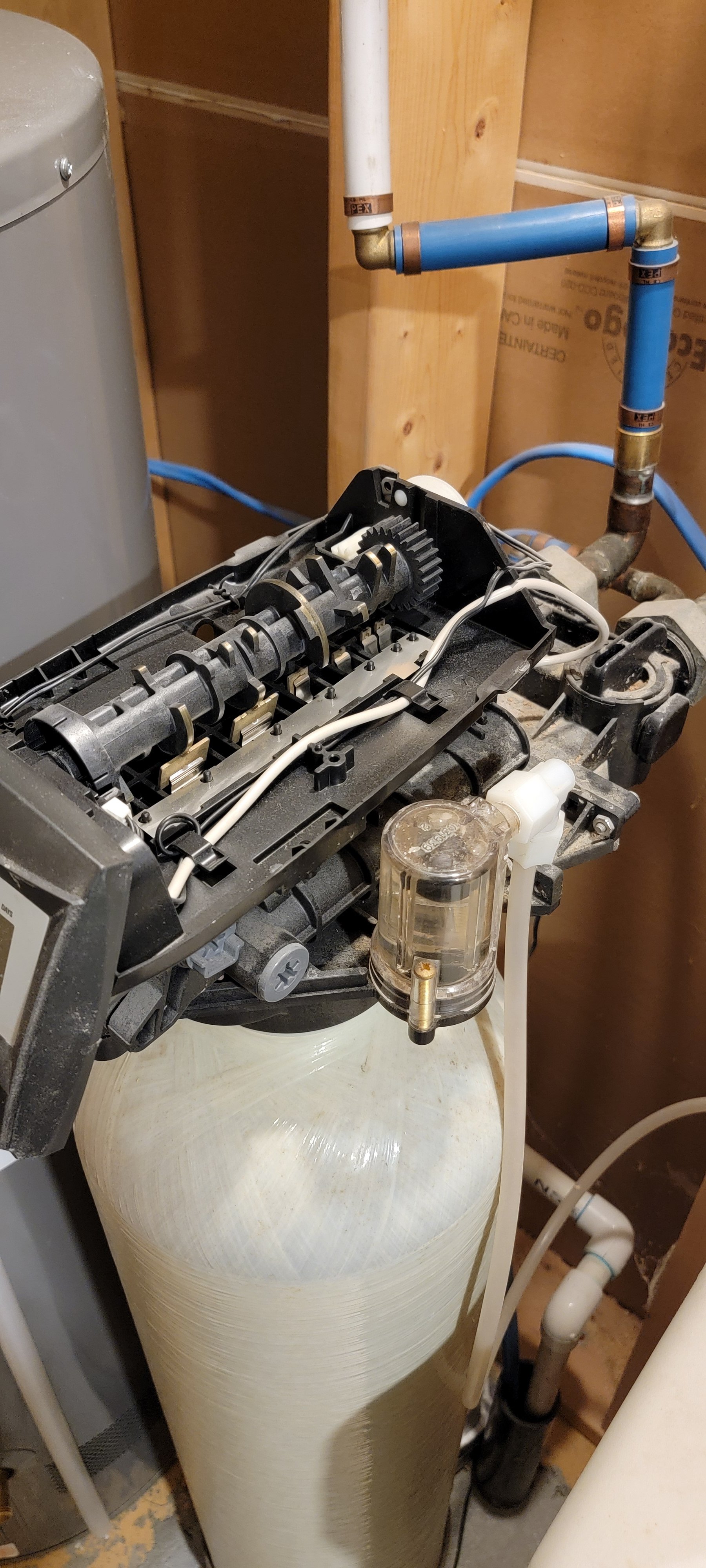 Exposed water-treatment equipment with blue PEX tubing, brass fittings, gears, wiring, and a large fiberglass tank.