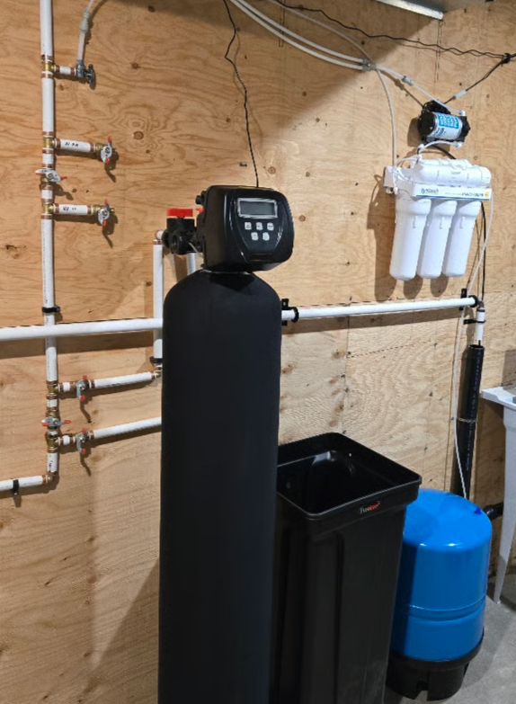 Water-treatment equipment with black and blue tanks, PVC pipes, valves, and a three-filter assembly on a plywood wall.