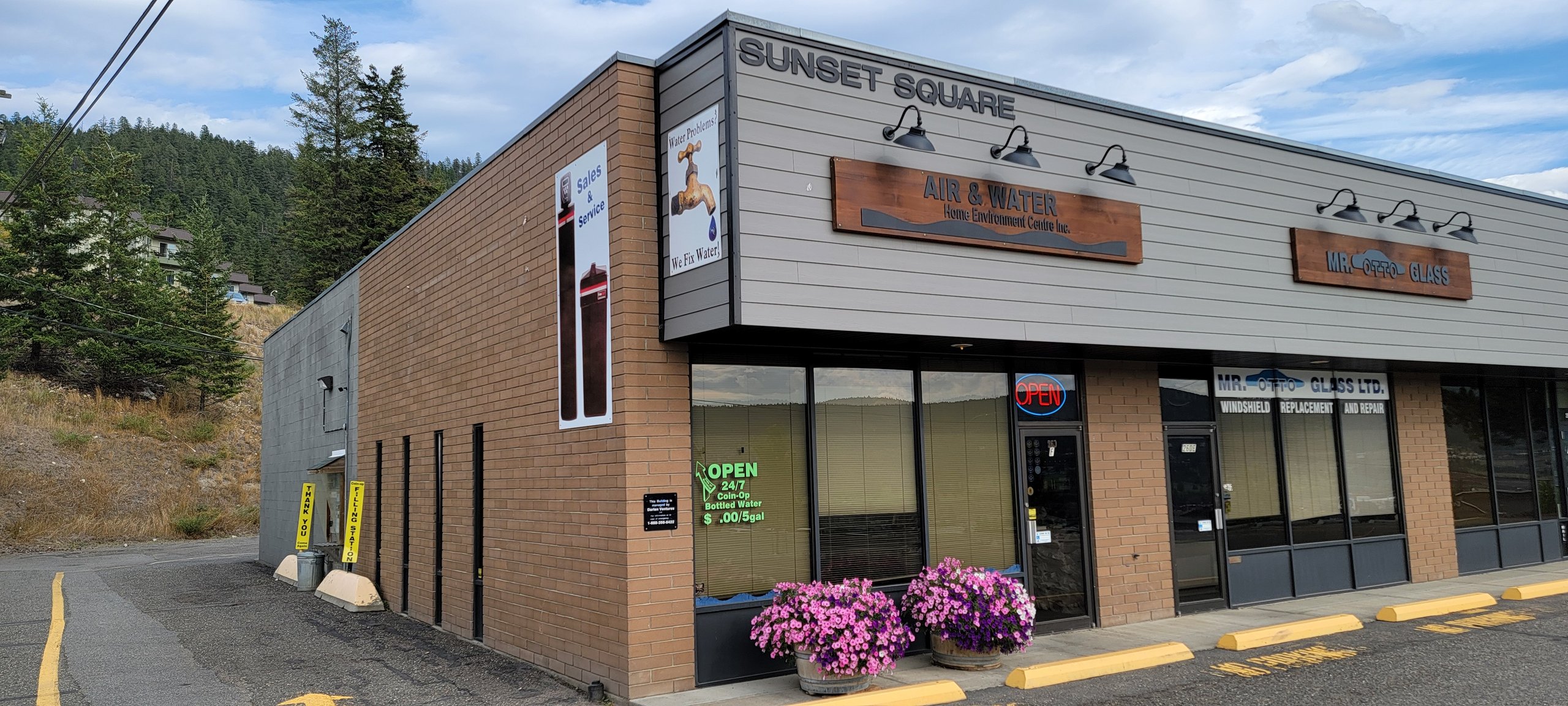Commercial storefront labeled Sunset Square with air, water, and auto glass signs beside a forested hillside.