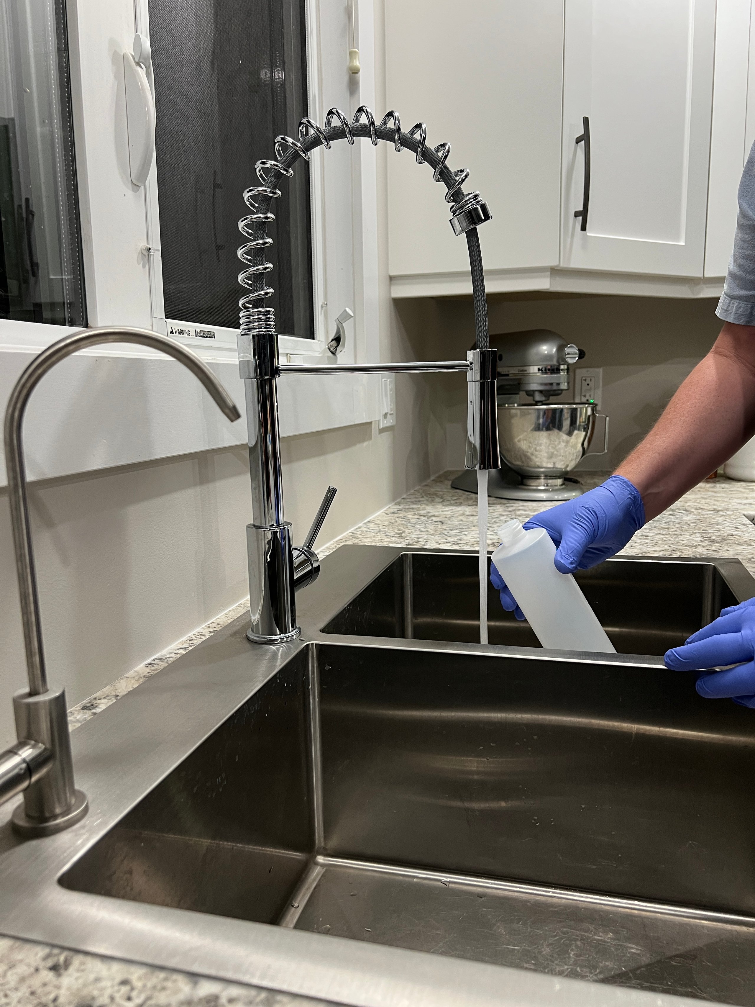 Gloved hands rinse a translucent bottle under a chrome pull-down faucet over a double kitchen sink.