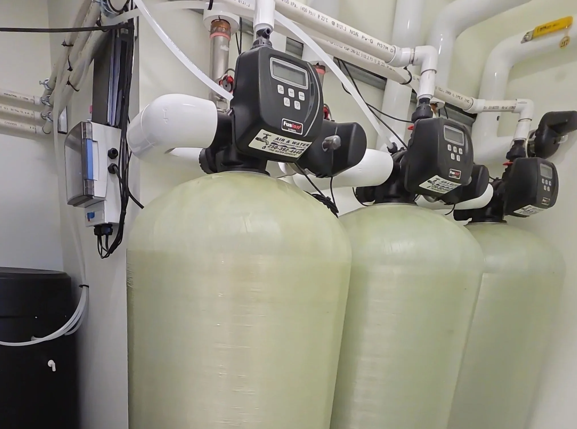 Three pale water-treatment tanks with black Fusion control heads, white PVC piping, and utility components.