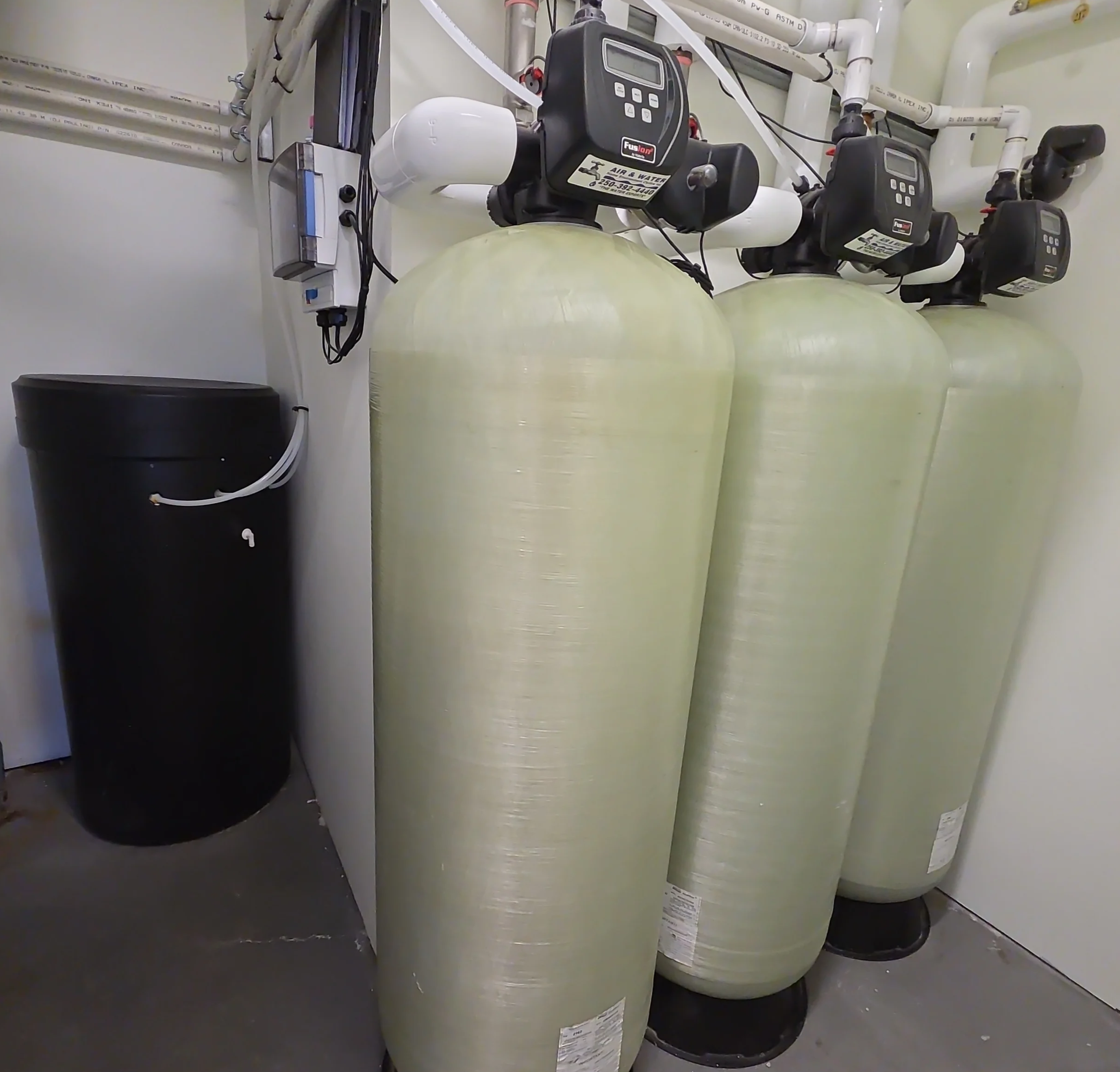 Three pale fiberglass water-treatment tanks with black Fusion control valves and white PVC plumbing in a utility room.