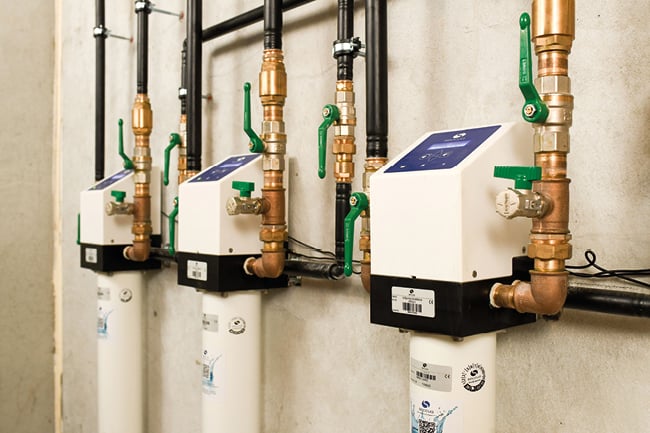 Three wall-mounted plumbing units with copper pipes, black tubing, green valve handles, and white cylindrical components.
